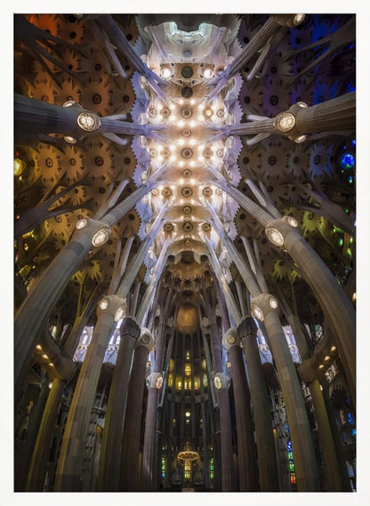 A low-angle, symmetrical photograph looking up at the incredibly ornate and complex vaulted ceiling of the Sagrada Familia Basilica. The massive stone columns branch out like trees, supporting the intricately detailed ceiling illuminated by warm, glowing lights and splashes of color from unseen stained glass windows. Poster