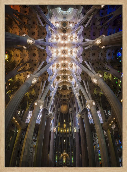 A low-angle, symmetrical photograph looking up at the incredibly ornate and complex vaulted ceiling of the Sagrada Familia Basilica. The massive stone columns branch out like trees, supporting the intricately detailed ceiling illuminated by warm, glowing lights and splashes of color from unseen stained glass windows. Poster