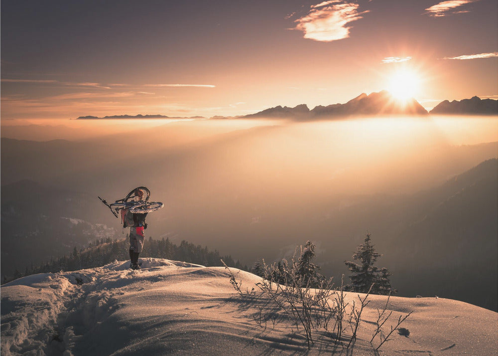 A lone person stands on a snowy mountain peak at sunrise, carrying a bicycle over their shoulder. They look out over a valley filled with a sea of clouds, with distant mountain ranges silhouetted against the bright, golden sun. Artwork