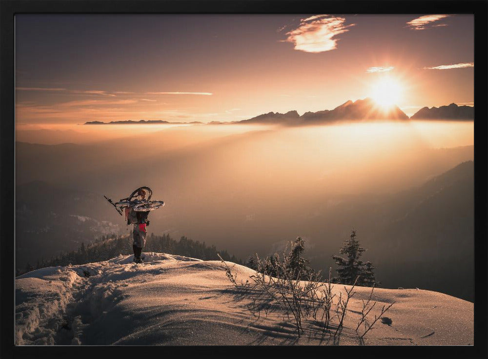 A lone person stands on a snowy mountain peak at sunrise, carrying a bicycle over their shoulder. They look out over a valley filled with a sea of clouds, with distant mountain ranges silhouetted against the bright, golden sun. Artwork