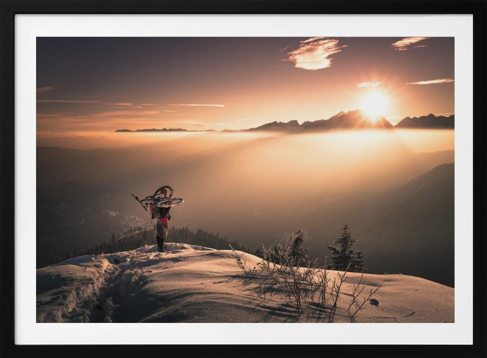A lone person stands on a snowy mountain peak at sunrise, carrying a bicycle over their shoulder. They look out over a valley filled with a sea of clouds, with distant mountain ranges silhouetted against the bright, golden sun. Artwork