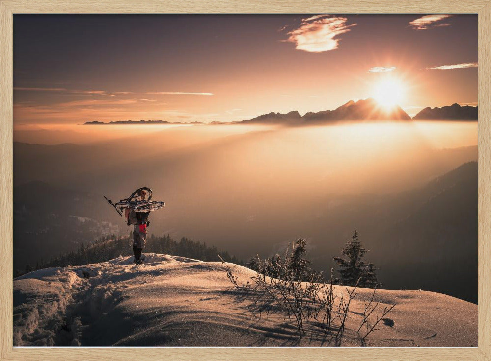 A lone person stands on a snowy mountain peak at sunrise, carrying a bicycle over their shoulder. They look out over a valley filled with a sea of clouds, with distant mountain ranges silhouetted against the bright, golden sun. Artwork