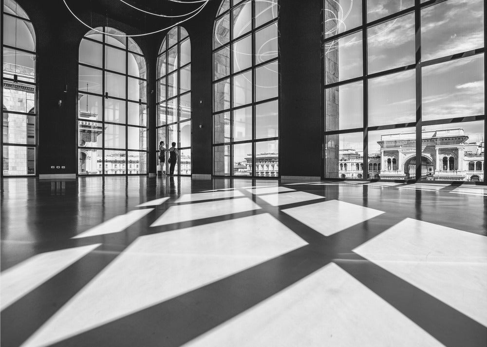 A striking black and white photograph of a spacious, modern hall with a polished floor. Dramatic shadows from large, arched windows are cast across the floor. Through the windows, a view of a classical European city with a triumphal arch can be seen. Print