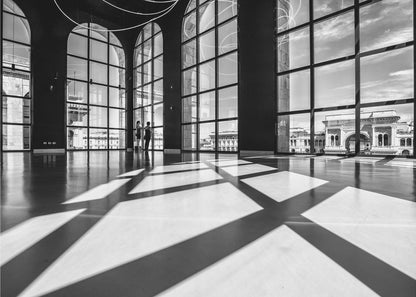 A striking black and white photograph of a spacious, modern hall with a polished floor. Dramatic shadows from large, arched windows are cast across the floor. Through the windows, a view of a classical European city with a triumphal arch can be seen. Print
