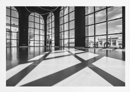 A striking black and white photograph of a spacious, modern hall with a polished floor. Dramatic shadows from large, arched windows are cast across the floor. Through the windows, a view of a classical European city with a triumphal arch can be seen. Print