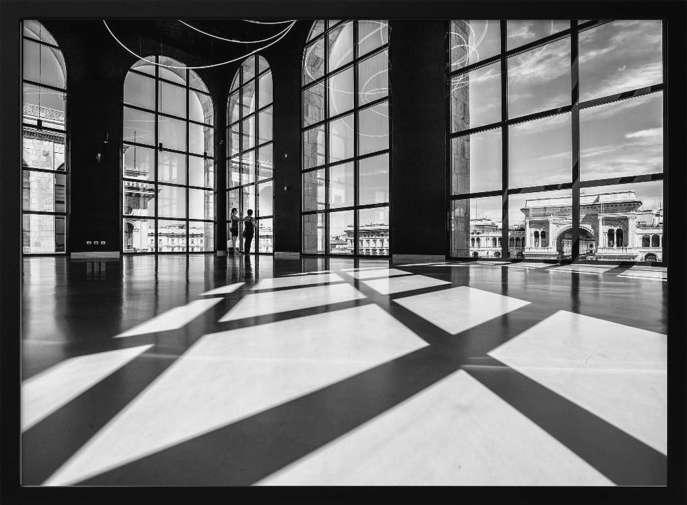 A striking black and white photograph of a spacious, modern hall with a polished floor. Dramatic shadows from large, arched windows are cast across the floor. Through the windows, a view of a classical European city with a triumphal arch can be seen. Print