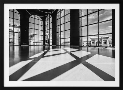 A striking black and white photograph of a spacious, modern hall with a polished floor. Dramatic shadows from large, arched windows are cast across the floor. Through the windows, a view of a classical European city with a triumphal arch can be seen. Print