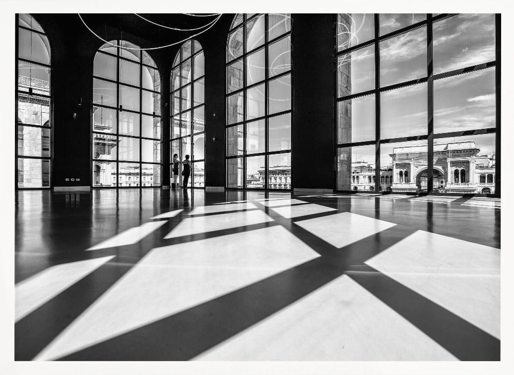 A striking black and white photograph of a spacious, modern hall with a polished floor. Dramatic shadows from large, arched windows are cast across the floor. Through the windows, a view of a classical European city with a triumphal arch can be seen. Print