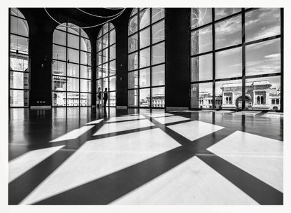A striking black and white photograph of a spacious, modern hall with a polished floor. Dramatic shadows from large, arched windows are cast across the floor. Through the windows, a view of a classical European city with a triumphal arch can be seen. Print