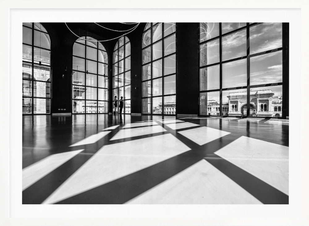A striking black and white photograph of a spacious, modern hall with a polished floor. Dramatic shadows from large, arched windows are cast across the floor. Through the windows, a view of a classical European city with a triumphal arch can be seen. Print