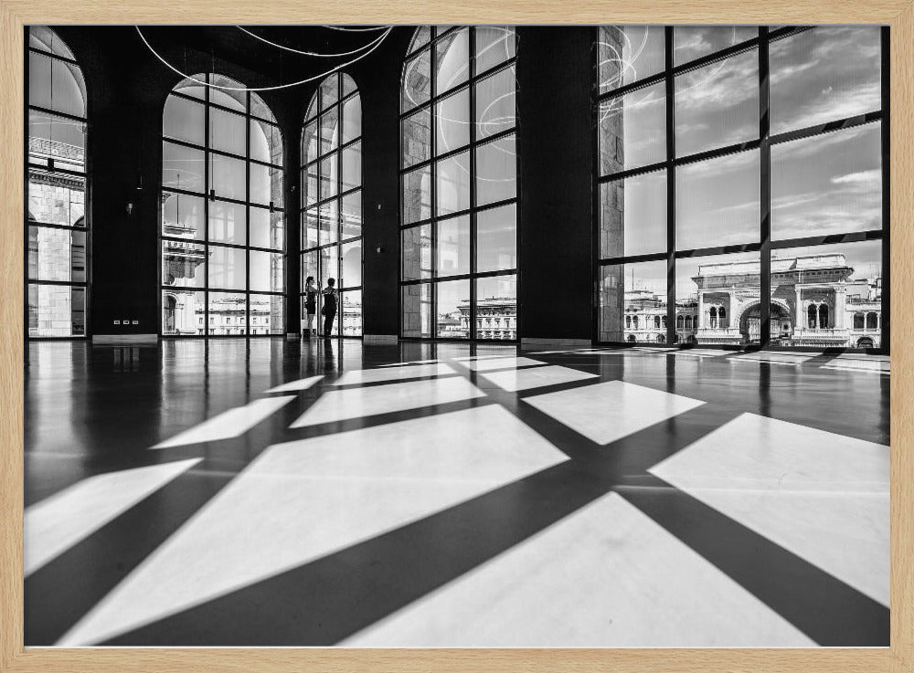 A striking black and white photograph of a spacious, modern hall with a polished floor. Dramatic shadows from large, arched windows are cast across the floor. Through the windows, a view of a classical European city with a triumphal arch can be seen. Print