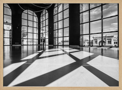 A striking black and white photograph of a spacious, modern hall with a polished floor. Dramatic shadows from large, arched windows are cast across the floor. Through the windows, a view of a classical European city with a triumphal arch can be seen. Print
