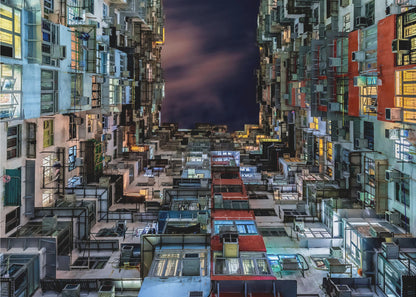 A low-angle nighttime view looking up from a courtyard surrounded by extremely dense, stacked apartment buildings with many windows lit in yellow, blue, and white, revealing a small patch of dark, cloudy sky. The image is presented within a brushed silver frame. Artwork