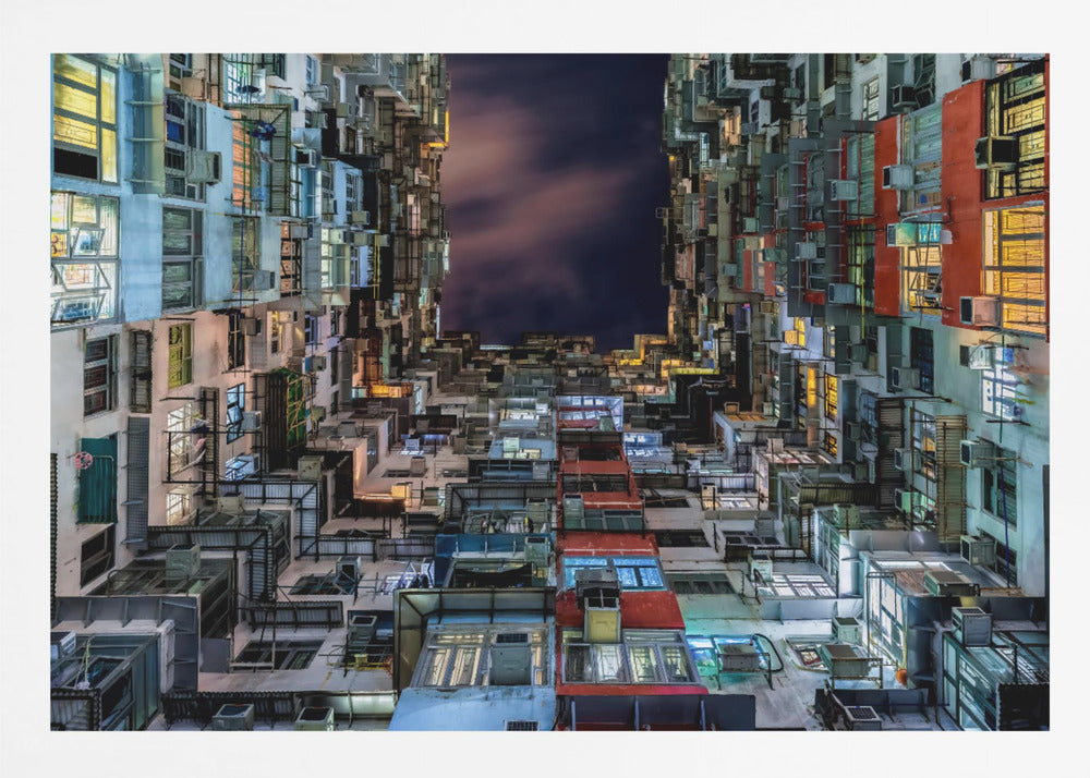 A low-angle nighttime view looking up from a courtyard surrounded by extremely dense, stacked apartment buildings with many windows lit in yellow, blue, and white, revealing a small patch of dark, cloudy sky. The image is presented within a brushed silver frame. Artwork