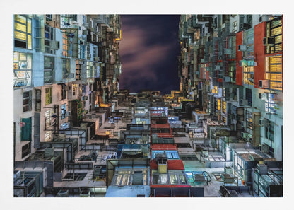 A low-angle nighttime view looking up from a courtyard surrounded by extremely dense, stacked apartment buildings with many windows lit in yellow, blue, and white, revealing a small patch of dark, cloudy sky. The image is presented within a brushed silver frame. Artwork