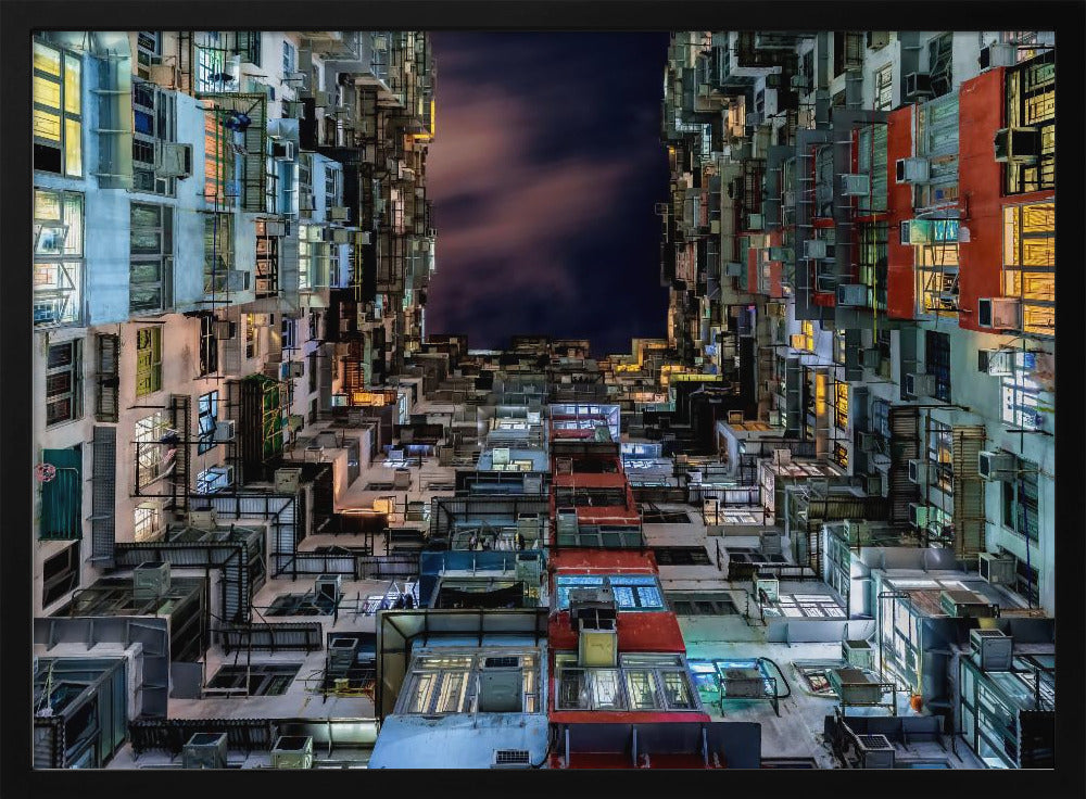 A low-angle nighttime view looking up from a courtyard surrounded by extremely dense, stacked apartment buildings with many windows lit in yellow, blue, and white, revealing a small patch of dark, cloudy sky. The image is presented within a brushed silver frame. Artwork