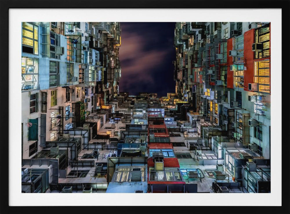 A low-angle nighttime view looking up from a courtyard surrounded by extremely dense, stacked apartment buildings with many windows lit in yellow, blue, and white, revealing a small patch of dark, cloudy sky. The image is presented within a brushed silver frame. Artwork