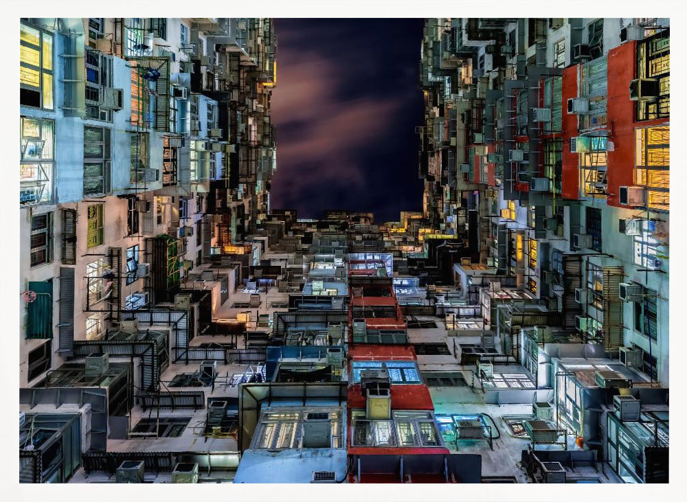 A low-angle nighttime view looking up from a courtyard surrounded by extremely dense, stacked apartment buildings with many windows lit in yellow, blue, and white, revealing a small patch of dark, cloudy sky. The image is presented within a brushed silver frame. Artwork