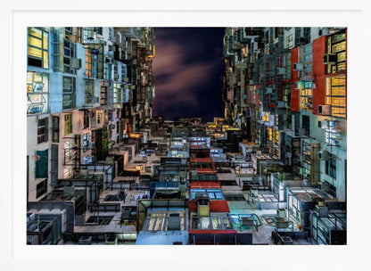 A low-angle nighttime view looking up from a courtyard surrounded by extremely dense, stacked apartment buildings with many windows lit in yellow, blue, and white, revealing a small patch of dark, cloudy sky. The image is presented within a brushed silver frame. Artwork