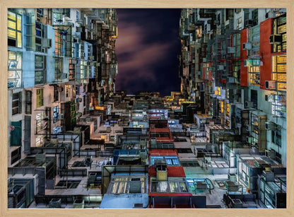 A low-angle nighttime view looking up from a courtyard surrounded by extremely dense, stacked apartment buildings with many windows lit in yellow, blue, and white, revealing a small patch of dark, cloudy sky. The image is presented within a brushed silver frame. Artwork