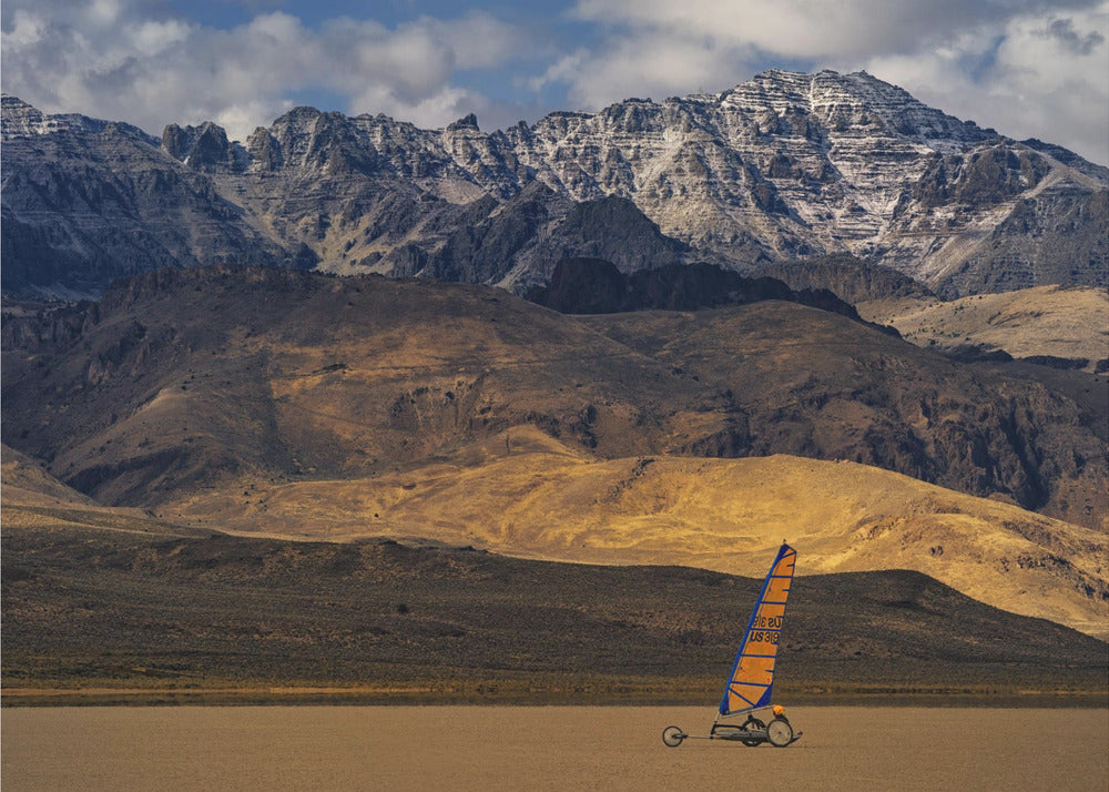A framed photograph of a land sailor with a bright orange and blue sail on a vast, flat desert floor. In the background, majestic snow-dusted mountains rise against a cloudy blue sky. Artwork