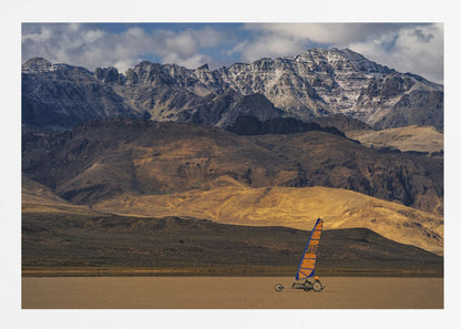 A framed photograph of a land sailor with a bright orange and blue sail on a vast, flat desert floor. In the background, majestic snow-dusted mountains rise against a cloudy blue sky. Artwork
