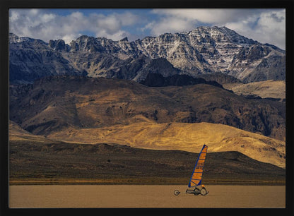 A framed photograph of a land sailor with a bright orange and blue sail on a vast, flat desert floor. In the background, majestic snow-dusted mountains rise against a cloudy blue sky. Artwork
