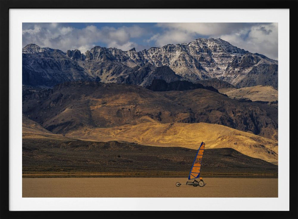 A framed photograph of a land sailor with a bright orange and blue sail on a vast, flat desert floor. In the background, majestic snow-dusted mountains rise against a cloudy blue sky. Artwork