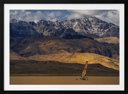 A framed photograph of a land sailor with a bright orange and blue sail on a vast, flat desert floor. In the background, majestic snow-dusted mountains rise against a cloudy blue sky. Artwork