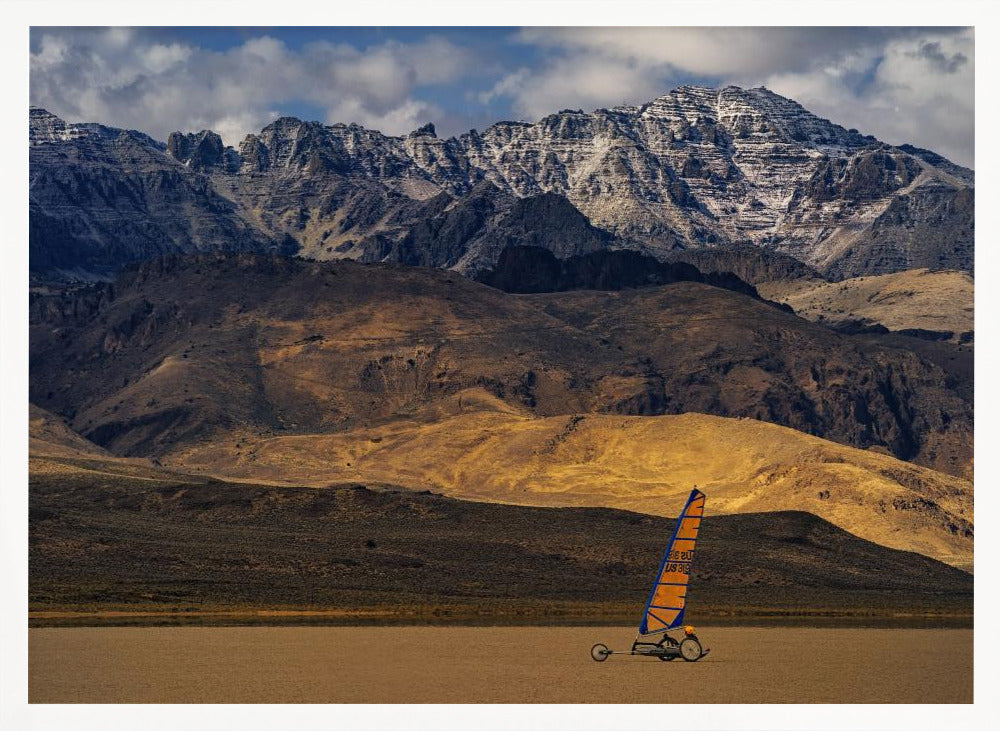 A framed photograph of a land sailor with a bright orange and blue sail on a vast, flat desert floor. In the background, majestic snow-dusted mountains rise against a cloudy blue sky. Artwork