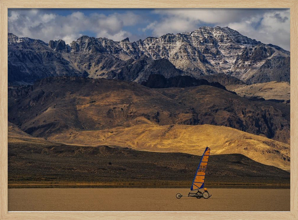 A framed photograph of a land sailor with a bright orange and blue sail on a vast, flat desert floor. In the background, majestic snow-dusted mountains rise against a cloudy blue sky. Artwork