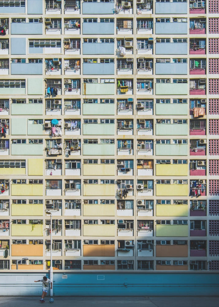 A symmetrical, full-frame shot of a massive apartment building with a repeating grid pattern of windows and balconies. The building is painted in horizontal bands of pastel colors, starting with blue at the top, then green, yellow, and orange. A single boy plays with a soccer ball on the ground level in front of a blue wall. Wall Art