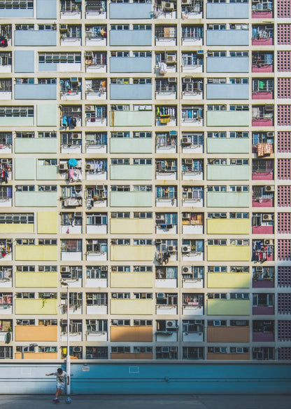 A symmetrical, full-frame shot of a massive apartment building with a repeating grid pattern of windows and balconies. The building is painted in horizontal bands of pastel colors, starting with blue at the top, then green, yellow, and orange. A single boy plays with a soccer ball on the ground level in front of a blue wall. Wall Art
