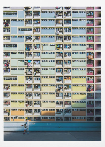 A symmetrical, full-frame shot of a massive apartment building with a repeating grid pattern of windows and balconies. The building is painted in horizontal bands of pastel colors, starting with blue at the top, then green, yellow, and orange. A single boy plays with a soccer ball on the ground level in front of a blue wall. Wall Art