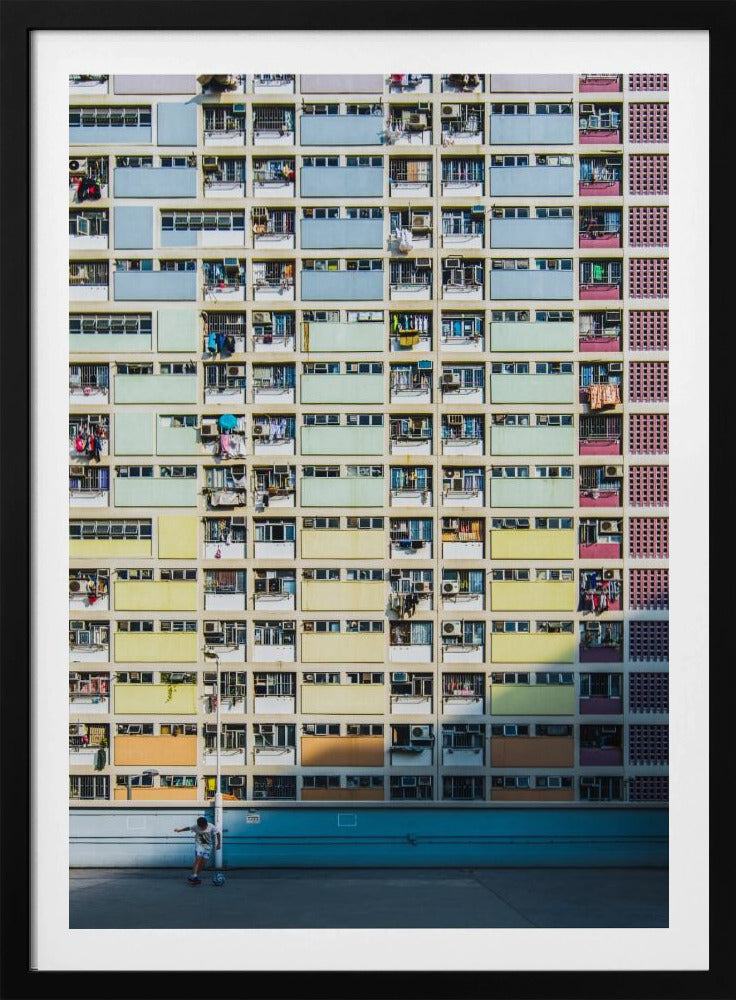 A symmetrical, full-frame shot of a massive apartment building with a repeating grid pattern of windows and balconies. The building is painted in horizontal bands of pastel colors, starting with blue at the top, then green, yellow, and orange. A single boy plays with a soccer ball on the ground level in front of a blue wall. Wall Art