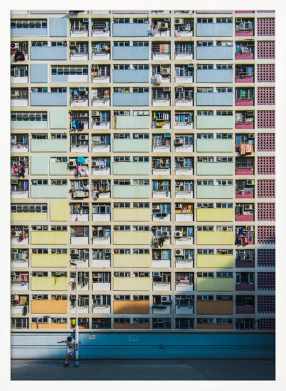 A symmetrical, full-frame shot of a massive apartment building with a repeating grid pattern of windows and balconies. The building is painted in horizontal bands of pastel colors, starting with blue at the top, then green, yellow, and orange. A single boy plays with a soccer ball on the ground level in front of a blue wall. Wall Art