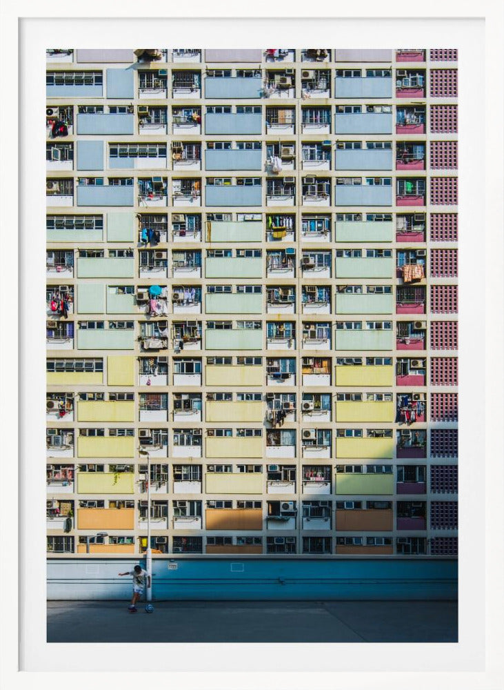 A symmetrical, full-frame shot of a massive apartment building with a repeating grid pattern of windows and balconies. The building is painted in horizontal bands of pastel colors, starting with blue at the top, then green, yellow, and orange. A single boy plays with a soccer ball on the ground level in front of a blue wall. Wall Art