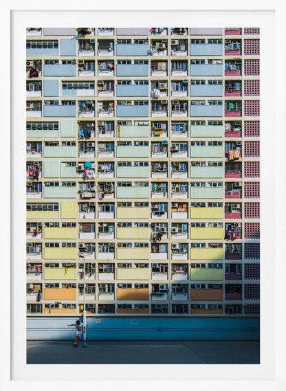 A symmetrical, full-frame shot of a massive apartment building with a repeating grid pattern of windows and balconies. The building is painted in horizontal bands of pastel colors, starting with blue at the top, then green, yellow, and orange. A single boy plays with a soccer ball on the ground level in front of a blue wall. Wall Art