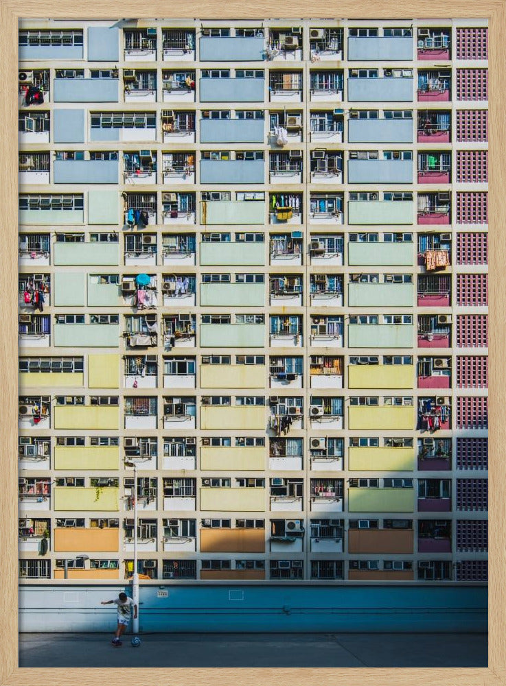 A symmetrical, full-frame shot of a massive apartment building with a repeating grid pattern of windows and balconies. The building is painted in horizontal bands of pastel colors, starting with blue at the top, then green, yellow, and orange. A single boy plays with a soccer ball on the ground level in front of a blue wall. Wall Art