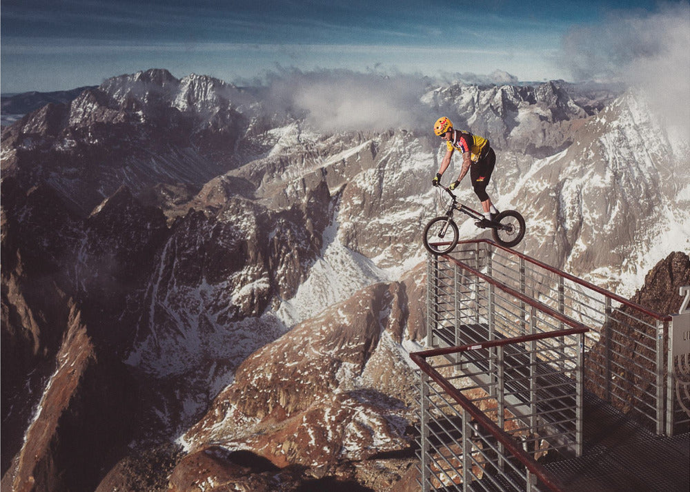 A trials cyclist wearing a yellow helmet balances precariously on the railing of a metal walkway high in the mountains, with a dramatic background of snow-dusted peaks and a blue sky. Poster
