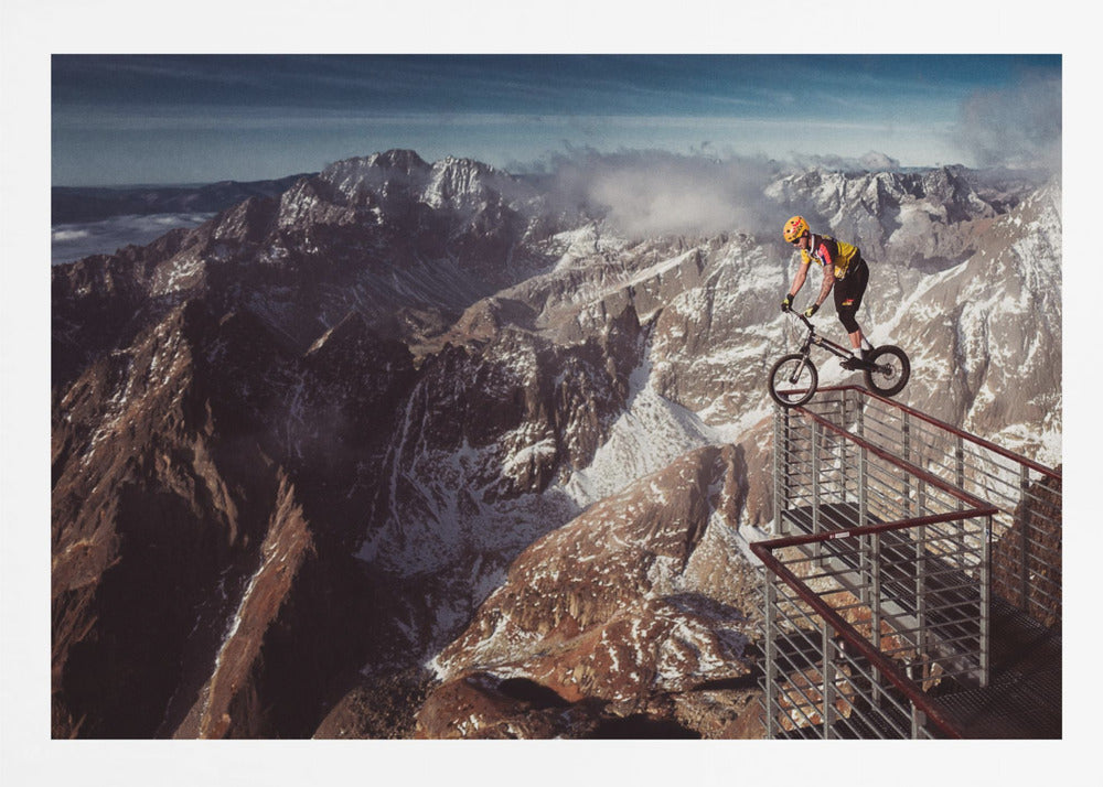 A trials cyclist wearing a yellow helmet balances precariously on the railing of a metal walkway high in the mountains, with a dramatic background of snow-dusted peaks and a blue sky. Poster
