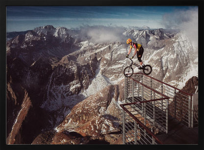 A trials cyclist wearing a yellow helmet balances precariously on the railing of a metal walkway high in the mountains, with a dramatic background of snow-dusted peaks and a blue sky. Poster