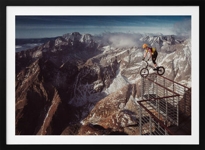 A trials cyclist wearing a yellow helmet balances precariously on the railing of a metal walkway high in the mountains, with a dramatic background of snow-dusted peaks and a blue sky. Poster