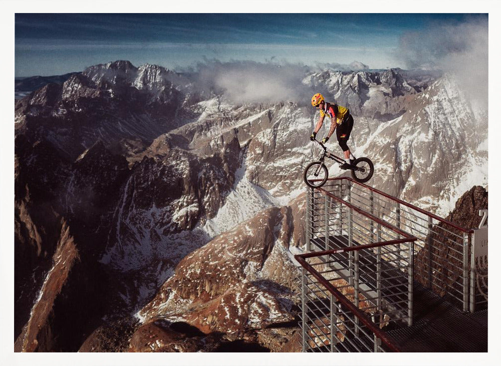 A trials cyclist wearing a yellow helmet balances precariously on the railing of a metal walkway high in the mountains, with a dramatic background of snow-dusted peaks and a blue sky. Poster