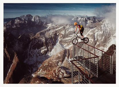 A trials cyclist wearing a yellow helmet balances precariously on the railing of a metal walkway high in the mountains, with a dramatic background of snow-dusted peaks and a blue sky. Poster