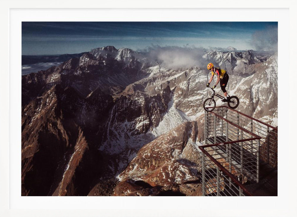 A trials cyclist wearing a yellow helmet balances precariously on the railing of a metal walkway high in the mountains, with a dramatic background of snow-dusted peaks and a blue sky. Poster