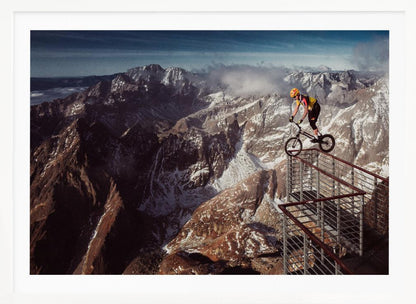 A trials cyclist wearing a yellow helmet balances precariously on the railing of a metal walkway high in the mountains, with a dramatic background of snow-dusted peaks and a blue sky. Poster