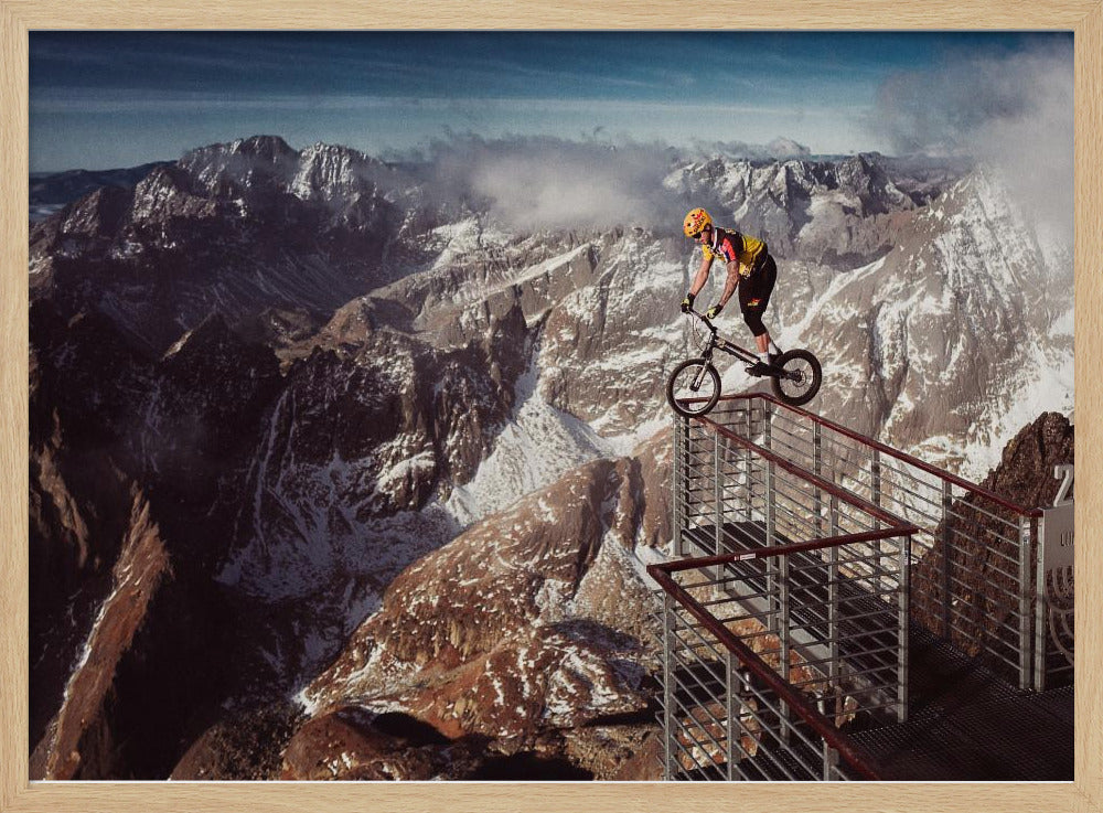 A trials cyclist wearing a yellow helmet balances precariously on the railing of a metal walkway high in the mountains, with a dramatic background of snow-dusted peaks and a blue sky. Poster
