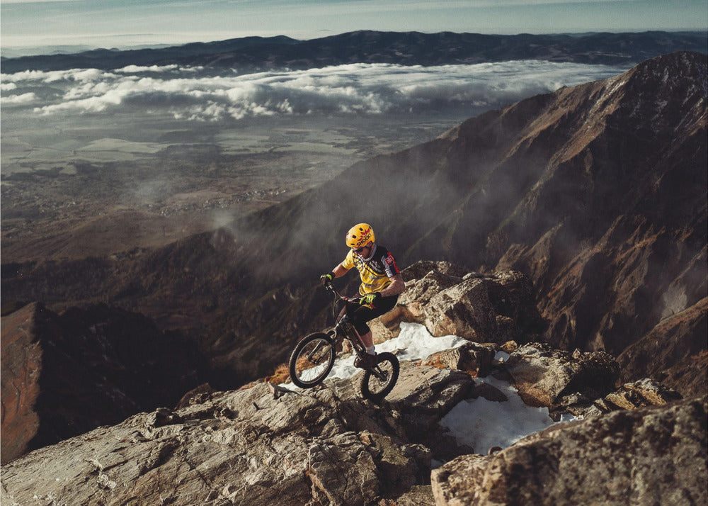 A mountain biker in a yellow helmet and jersey navigates a treacherous, rocky mountain ridge. Below, a vast valley is filled with a sea of clouds, with distant mountain ranges visible on the horizon. Print