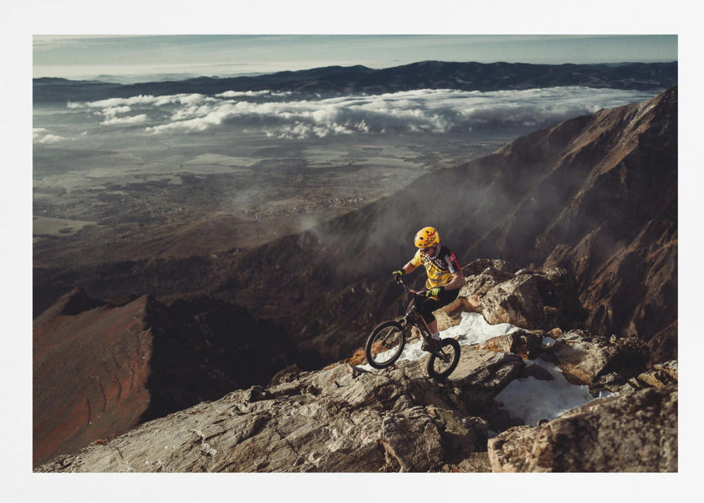 A mountain biker in a yellow helmet and jersey navigates a treacherous, rocky mountain ridge. Below, a vast valley is filled with a sea of clouds, with distant mountain ranges visible on the horizon. Print